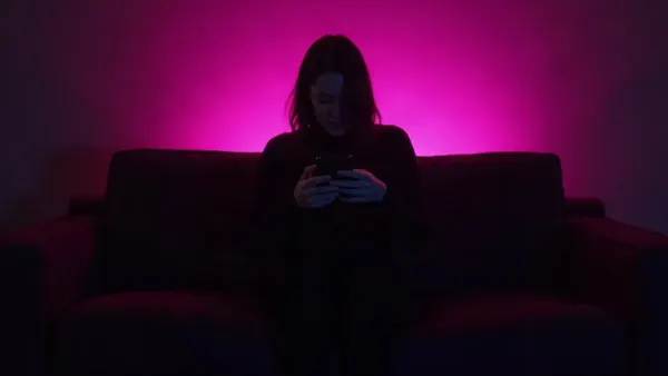 Silhouette di spalle su un divano con un telefono in mano, in una stanza buia dal bagliore magenta (illustrazione dell’articolo)